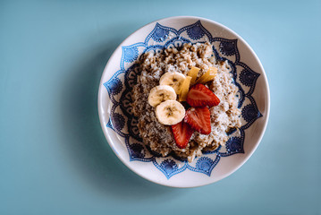 Oatmeal with strawberries, banana honey and coconut flakes in a plate with a blue ornament. Healthy organic food. Tasty breakfast on a blue background. Soft focus