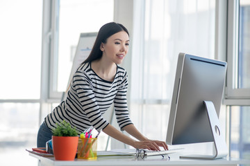 Dark-haired woman in a striped shirt standing near the table and looking at the computer
