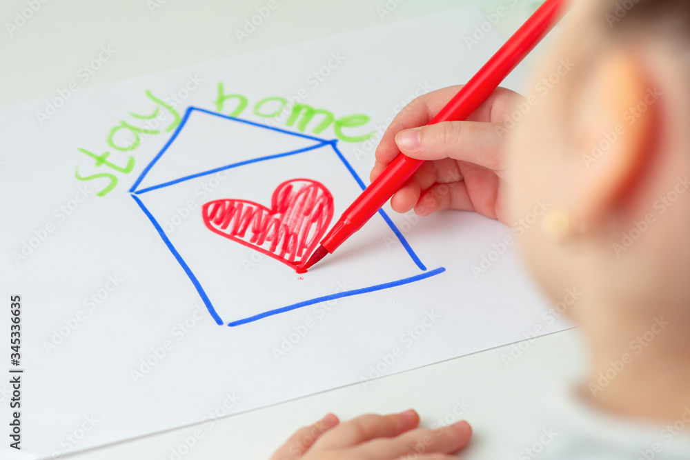 Wall mural Stay Home concept. Child is drawing house with red heart on the sheet of paper with a written phrase over the house Stay Home on the easel.