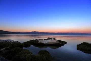 Sundown over the lake and big mossy stones in the water, mountains on the horizon -- Sunset seascape, background. Lake Sevan. Armenia. Сoncept of tranquility of a pacification