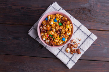 Fresh caramel popcorn and colorful candies mix in bowl on white napkin. Brown wooden table from top view.