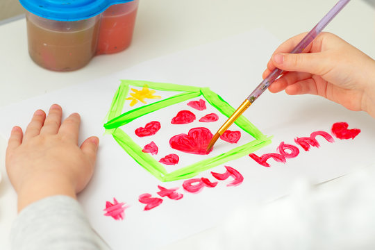 Child is drawing heart inside house on white sheet of paper with words Stay Home. Stay Home concept. Children creativity.