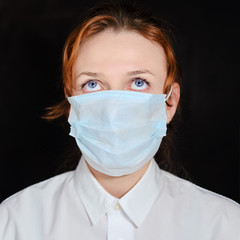 Redhead woman in a medical mask who is looking up in thought. The girl raised her eyes up on a black background. Concept of challenges with the coronavirus epidemic