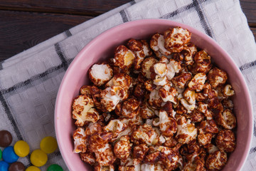 Close up salty popcorn covered chocolate in pink bowl and colorful candies on wooden background. Flat lay composition.