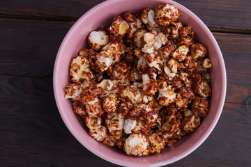 Close up delicious popcorn covered chocolate in pink bowl on wooden background. Homemade chocolate popcorn. Concept of watching tv at home.