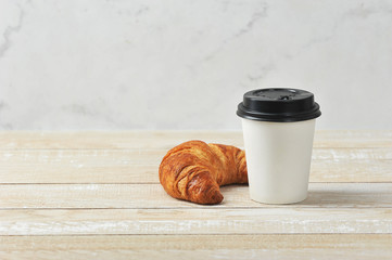 Croissant and a cup of coffee. Takeaway coffee and takeaway concept. Wooden light background.  Free space for text.