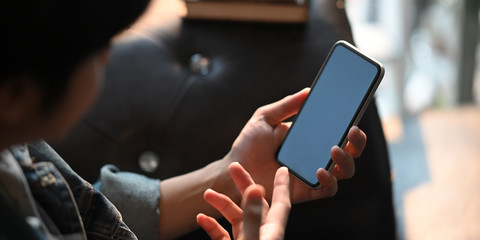 Cropped image of smart man hands holding and using a white blank screen smartphone while sitting and relaxing at the black leather sofa over comfortable sitting room as background.