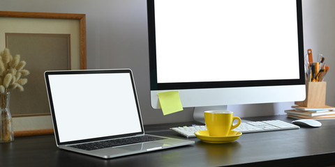 Workspace laptop with white blank screen putting on black working desk surrounded by wild grass in glass vase, empty picture frame, computer monitor with white screen, coffee cup and keyboard.