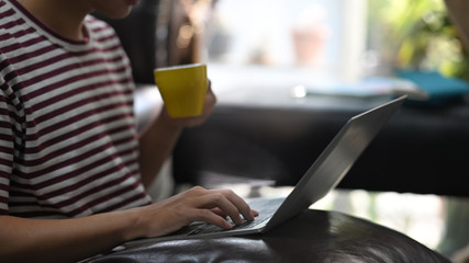 Cropped image of creative man holding a hot coffee cup while typing on computer laptop that putting on his lap and sitting at the leather couch over comfortable living room as background.