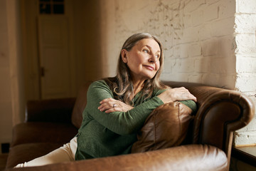 Indoor image of beautiful pensive sixty year old female with gray hair sitting comfortably in stylish leather sofa by window, having thoughtful dreamy facial expression, wants to have walk outside