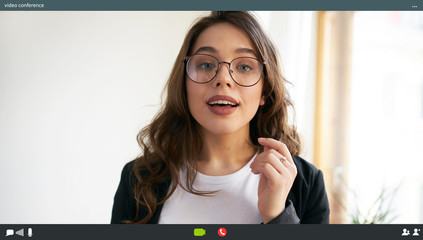 Indoor shot of energetic attractive young female manager in eyeglasses talking to colleagues online...