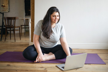 Beautiful happy young female with curvy body sitting on mat in front of open laptop, surfing...