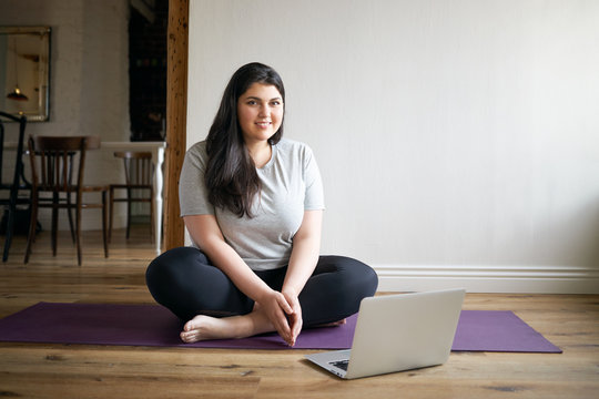 Cute Girl In Plus Size Sports Clothes Watching Online Video Tutorial By Fitness Instructor Via Portable Computer, Doing Exercises On Mat, Having Happy Look. Technology, Health And Activity Concept