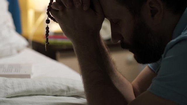 Man is praying. Bearded man sits on floor in bedroom by bed, holds rosary with crucifix in his hands and says prayer. Close up