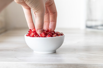 Hand takes pomegranate seeds from a small white bowl on a beige background