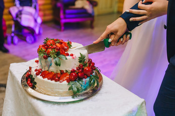 Newlyweds cut a wedding cream cake decorated with strawberry and cherry fruits, a cake at a party. Photography, concept.