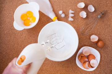 Female hands whisk the whites with a white bowl with a hand-held electric mixer on a wooden table. Step by step recipe of meringue cookies. Step 2. Top view.
