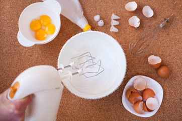 Female hands whisk the whites with a white bowl with a hand-held electric mixer on a wooden table. Step by step recipe of meringue cookies. Step 2. Top view.