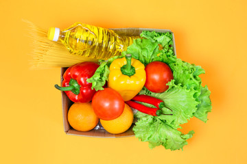 Cardboard box with food, sunflower oil, red, yellow pepper, chili, oranges, tomatoes, pasta, isolated over a yellow background, top view, donation, safe food delivery to your home, volunteer help