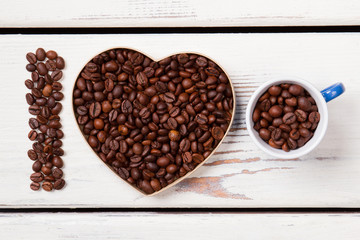 Heart shape form and cup filled with roasted coffee grains. I love natural fresh coffee. White wooden planks on background.