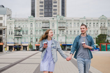 Romantic and happy caucasian couple in casual clothes walking together through the streets. Love, relationships, romance, happiness concept. Man and woman with disposable cups of coffee in the city.