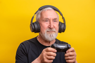 Portrait senior man with gray beard in wireless headphone holds game controller over yellow background with copy space