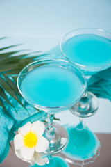 Exotic tropical Blue Curacao cocktail drink in a glasses with Plumeria frangipani flower, palm leaf on the background.
