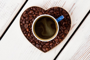 Cup of coffee standing over heart made of coffee grains. Coffee love concept. White wooden background.