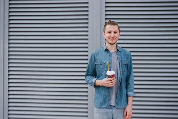 Young smiling caucasian man in casual clothes with braces drinking coffee in the city streets from disposable cup on urban background.