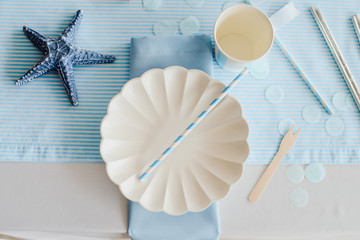 Tableware and decorations for serving festive table in sea style. Elegant plates, paper cups, straws with blue decorative textile. Birthday or baby shower boy .