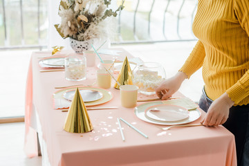 Decorator near party table in pastel colors with pink tablecloth, paper colorful dishes, cups and golden cutlery. Girl birthday party decoration, festive caps.