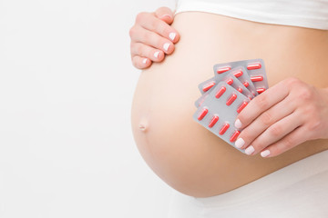 Young woman hand holding blister packs of red pills for improve of iron level in blood. Receiving vitamins in pregnancy time. Big naked belly. Side view. Close up. Isolated on light gray background.