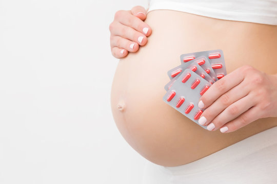 Young Woman Hand Holding Blister Packs Of Red Pills For Improve Of Iron Level In Blood. Receiving Vitamins In Pregnancy Time. Big Naked Belly. Side View. Close Up. Isolated On Light Gray Background.