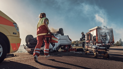 Paramedics and Firefighters Arrive On the Car Crash Traffic Accident Scene. Professionals Rescue Injured Victim Trapped in Rollover Vehicle by Extricating Them, giving First Aid and Extinguishing Fire