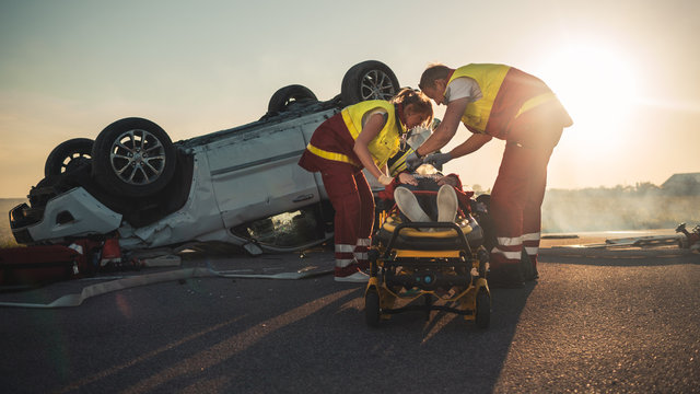 On the Car Crash Traffic Accident Scene: Paramedics Saving Life of a Female Victim who is Lying on Stretchers. They Listen To a Heartbeat, Apply Oxygen Mask and Give First Aid. Background Firefighters