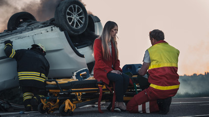 On the Car Crash Traffic Accident Scene: Paramedics Perform First Aid of a Female Victim who is Sitting on Stretchers. Rollover in the Background.