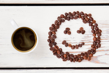 Top view cup of hot coffee and beans in a shape of smiley face. White wooden planks on background.