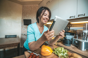 Senior woman cooking vegetables at home reading a recipe on the tablet. Old people using technology...