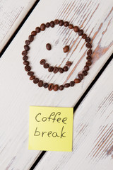 Smiley made of beans and sticker with coffee break writing. White wooden table surface on background.