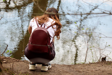 Redhead girl with a bag-backpack is squatting on the river bank. Outdoor recreation in spring