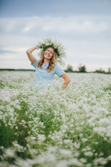 Naklejka na ściany i meble beautiful blonde girl in a field of daisies. wreath of wildflowers on his head. woman in a blue dress in a field of white flowers. charming girl with a bouquet of daisies. summer tender photo