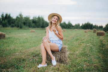 beautiful girl in a straw hat on a field with straw bales. girl sitting on the hay. blonde in the summer in a field with a hat. life in the countryside, outdoors. eco project. clean environment