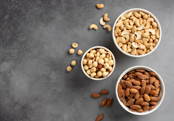 flat lay of various types of nuts (cashews, hazelnuts, almonds) in white bowls on a gray concrete background. food border. copy space.