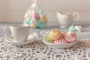 Multi-colored meringues in pastel shades with cups and a jug on a light textured wooden background. The concept of Breakfast with tea or coffee, home comfort and tenderness. Close-up, selective focus