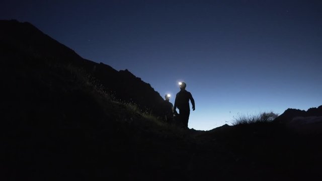 Silhouette of two men with head lamps hiking in the dark with mountain panorama, static, 4K