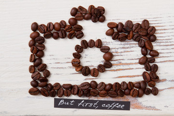 Roasted coffee beans arranged in a shape of photo camera. White wood on background. But first coffee.