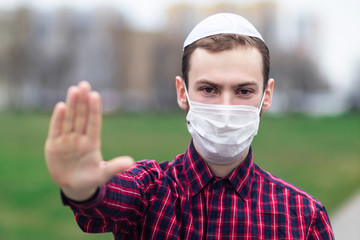 Handsome young Jewish guy in traditional jewish male headdress, hat, boom, or yiddish on head. Man in medical mask on his face showing palm, stop sign against coronavirus, virus pandemic. Covid-19