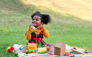Adorable African girl enjoys to playing her toys in park or garden during a picnic with her family.