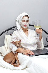 Woman taking care of her face with a face mask in bed, with dog, drinking a glass of wine