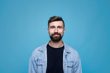 Young cheerful man in black t-shirt and denim jacket. Guy with beard expresses happiness. Blue background.